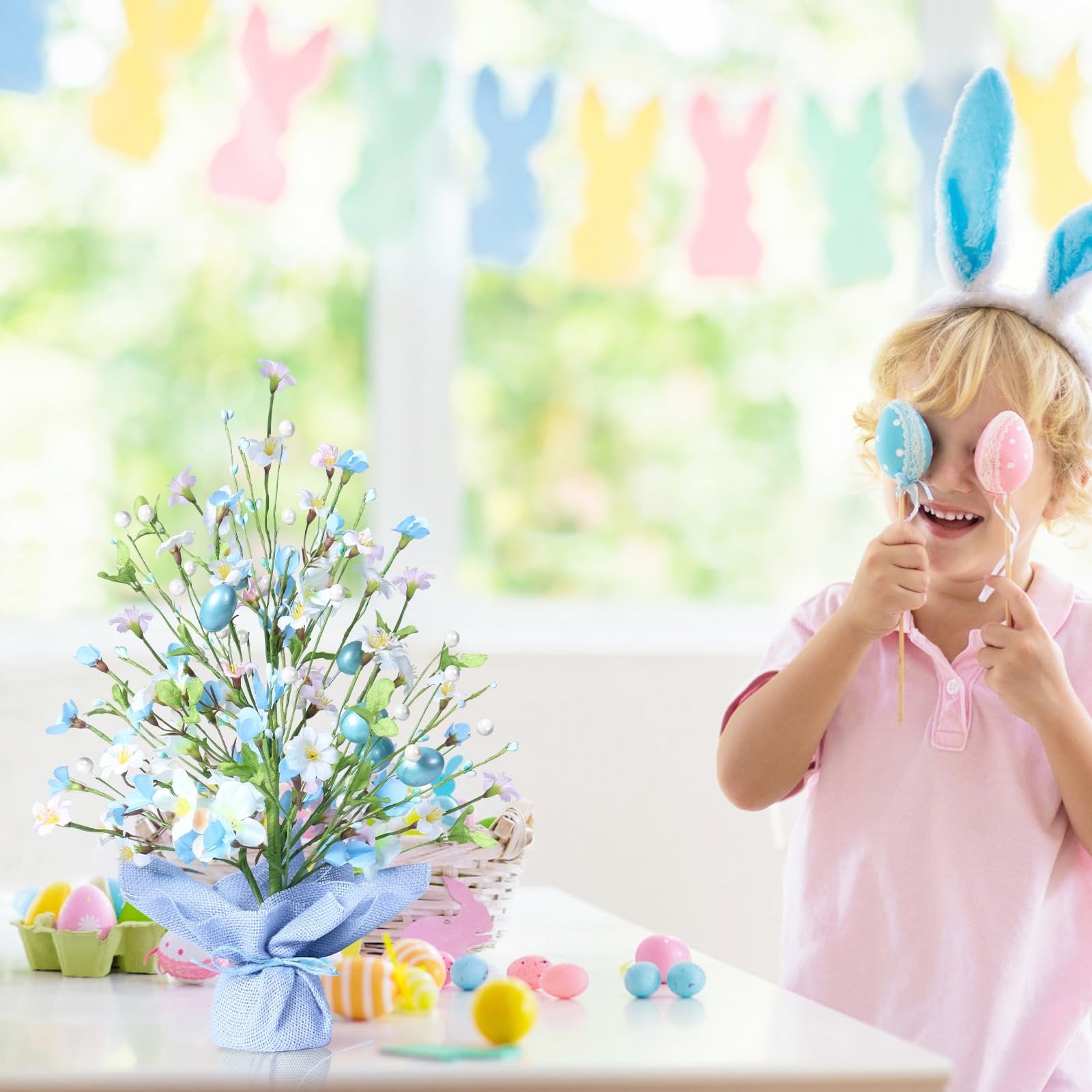 Easter tabletop tree with blue eggs and flowers, a child with bunny ears holds eggs to their eyes.