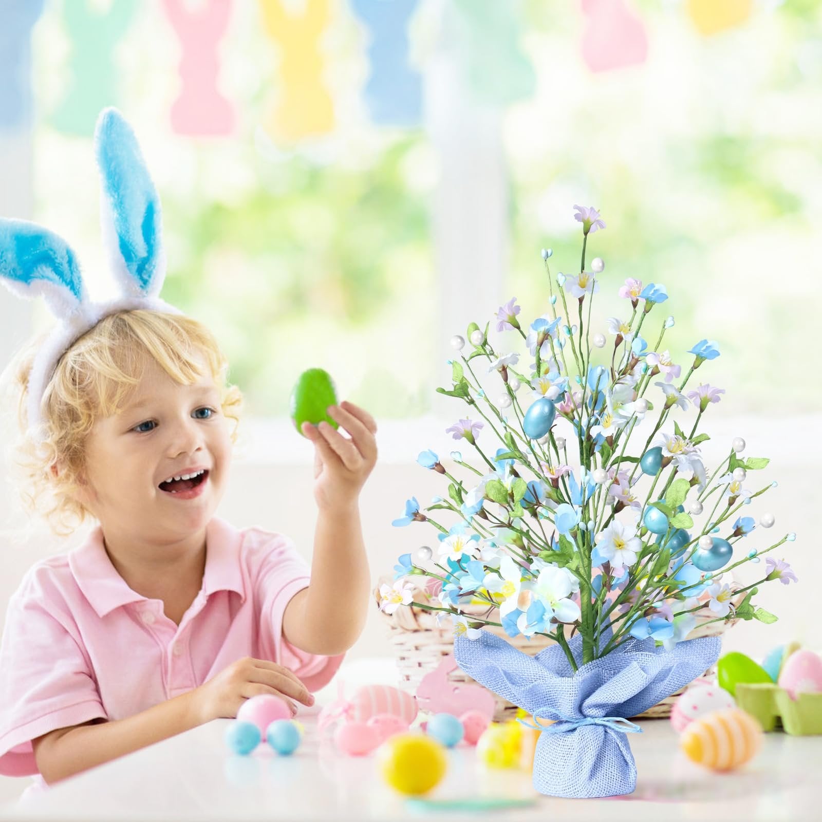 Young boy with bunny ears holding an Easter egg near a decorative Easter egg tree with blue eggs and flowers.