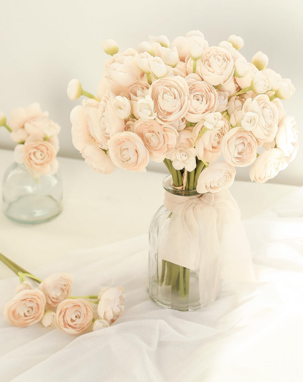 Champagne faux silk ranunculus flowers in a glass vase with a ribbon, and loose flowers on white fabric.