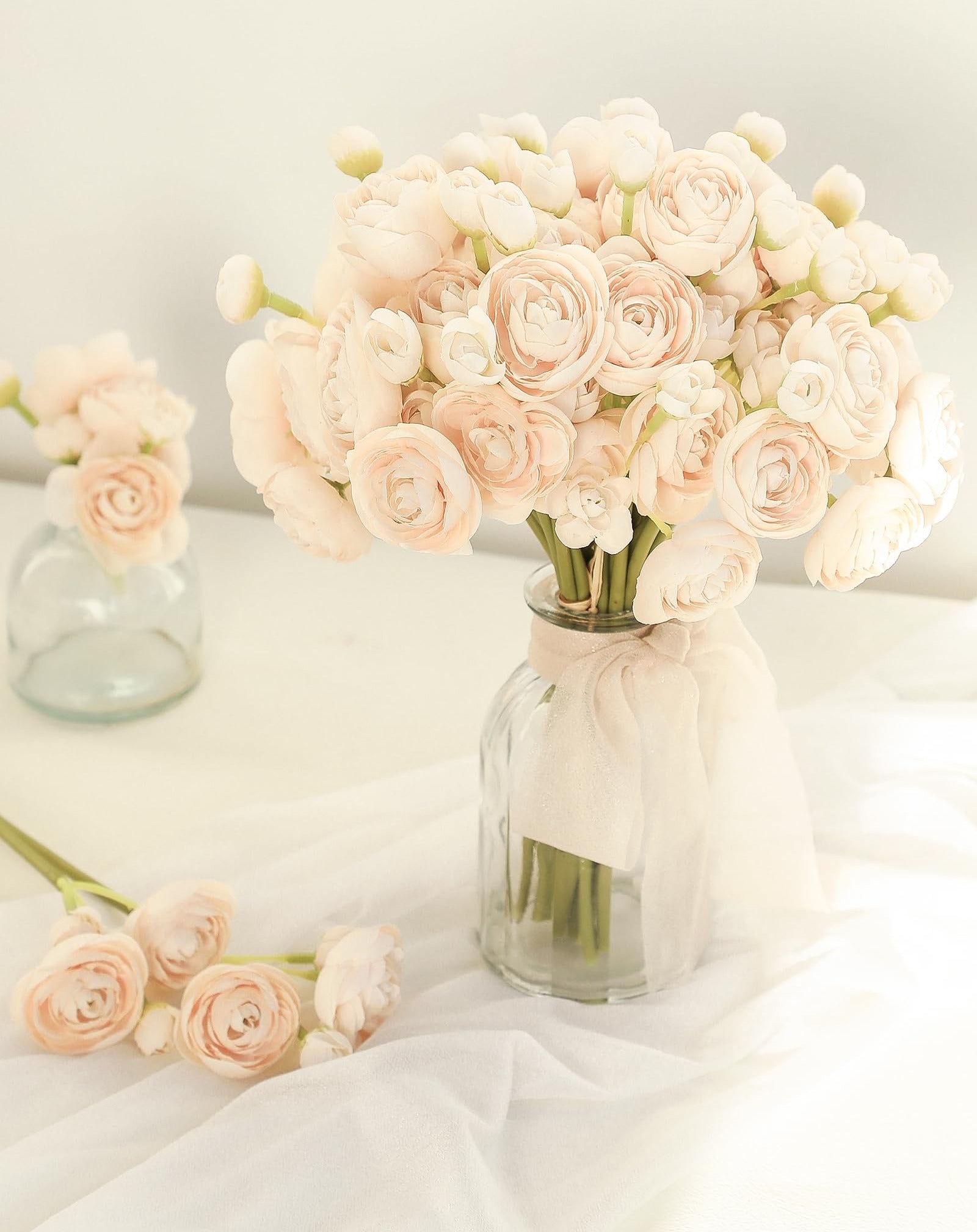 Champagne faux silk ranunculus flowers in a glass vase with a ribbon, and loose flowers on white fabric.