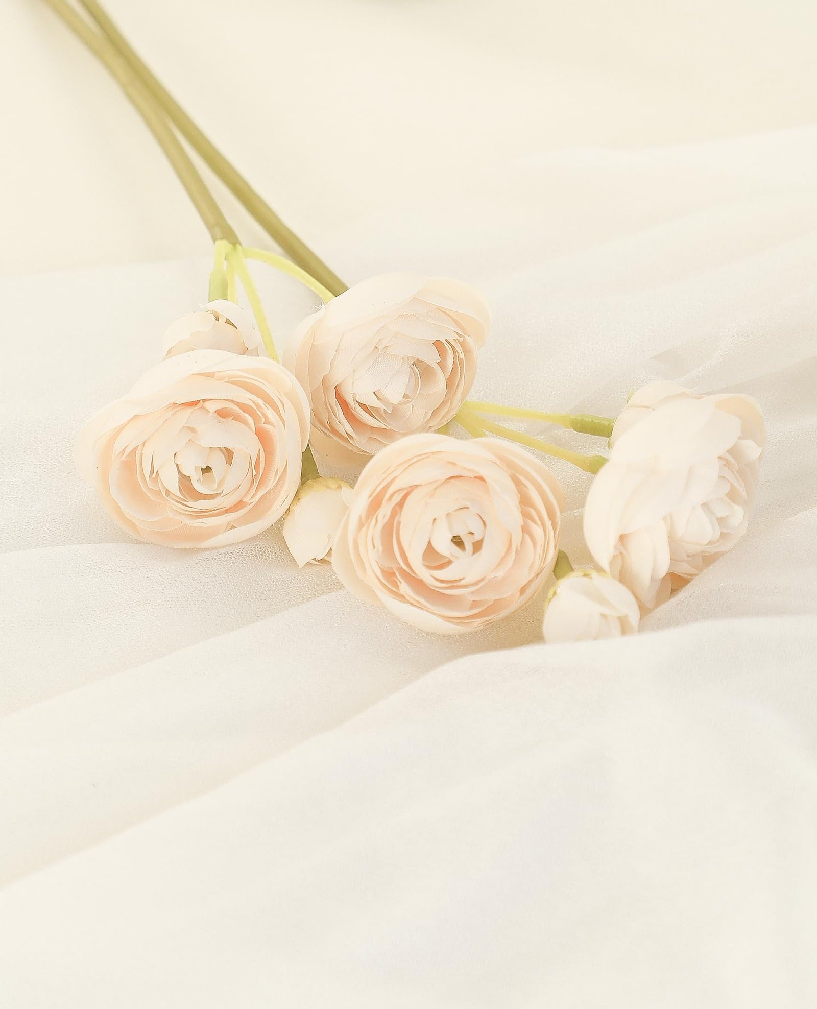 Champagne faux silk ranunculus flowers on a draped ivory fabric, with a blurred glass vase in the background.