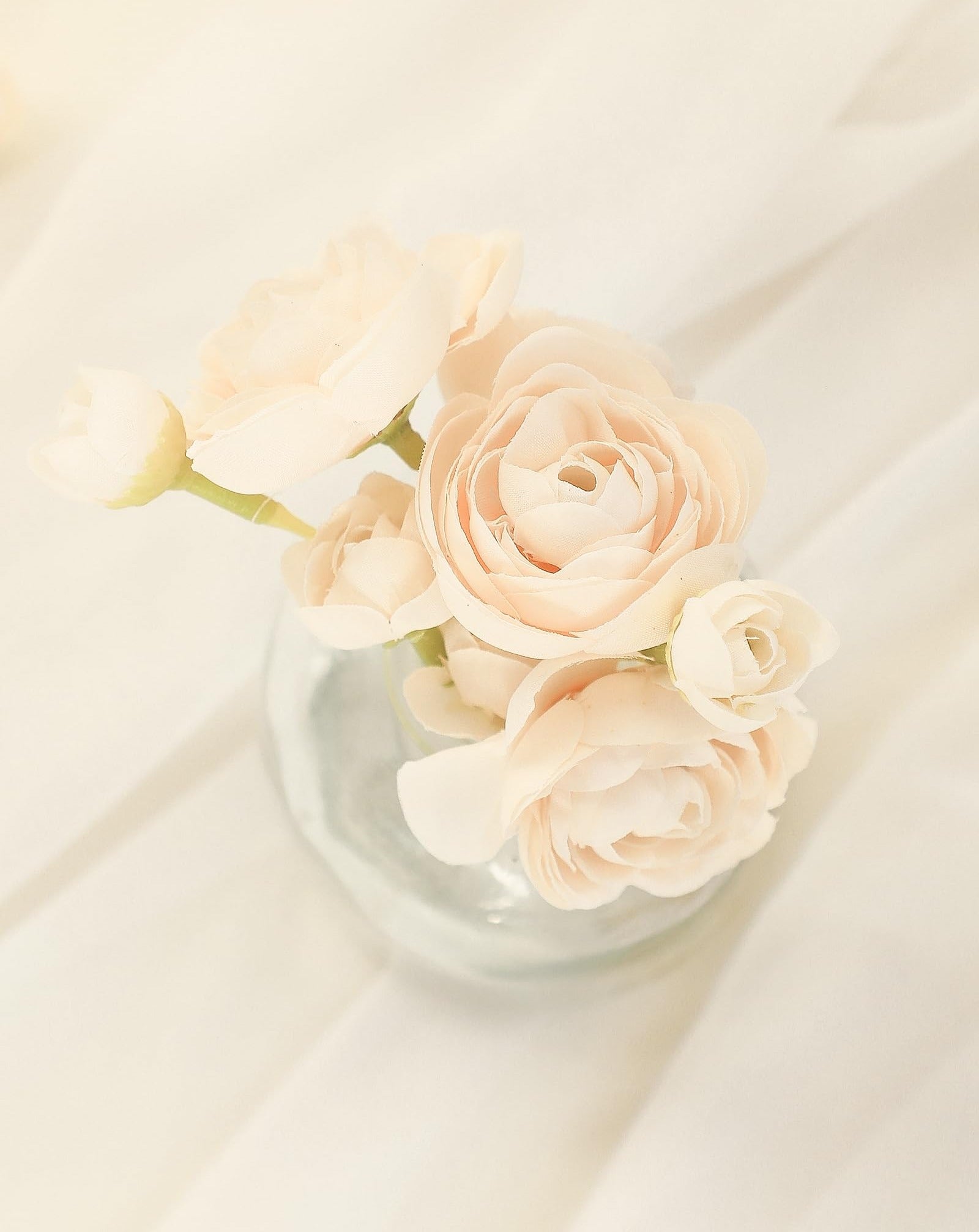 Champagne faux silk ranunculus flowers in a clear glass vase on a draped fabric background