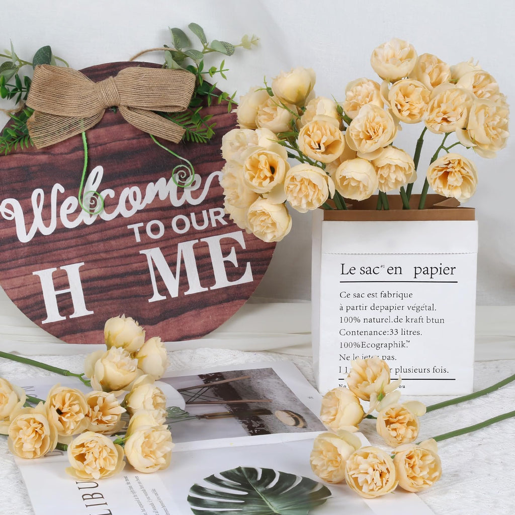 Champagne artificial peonies and ranunculus flowers arranged with a wooden welcome sign and a paper bag.