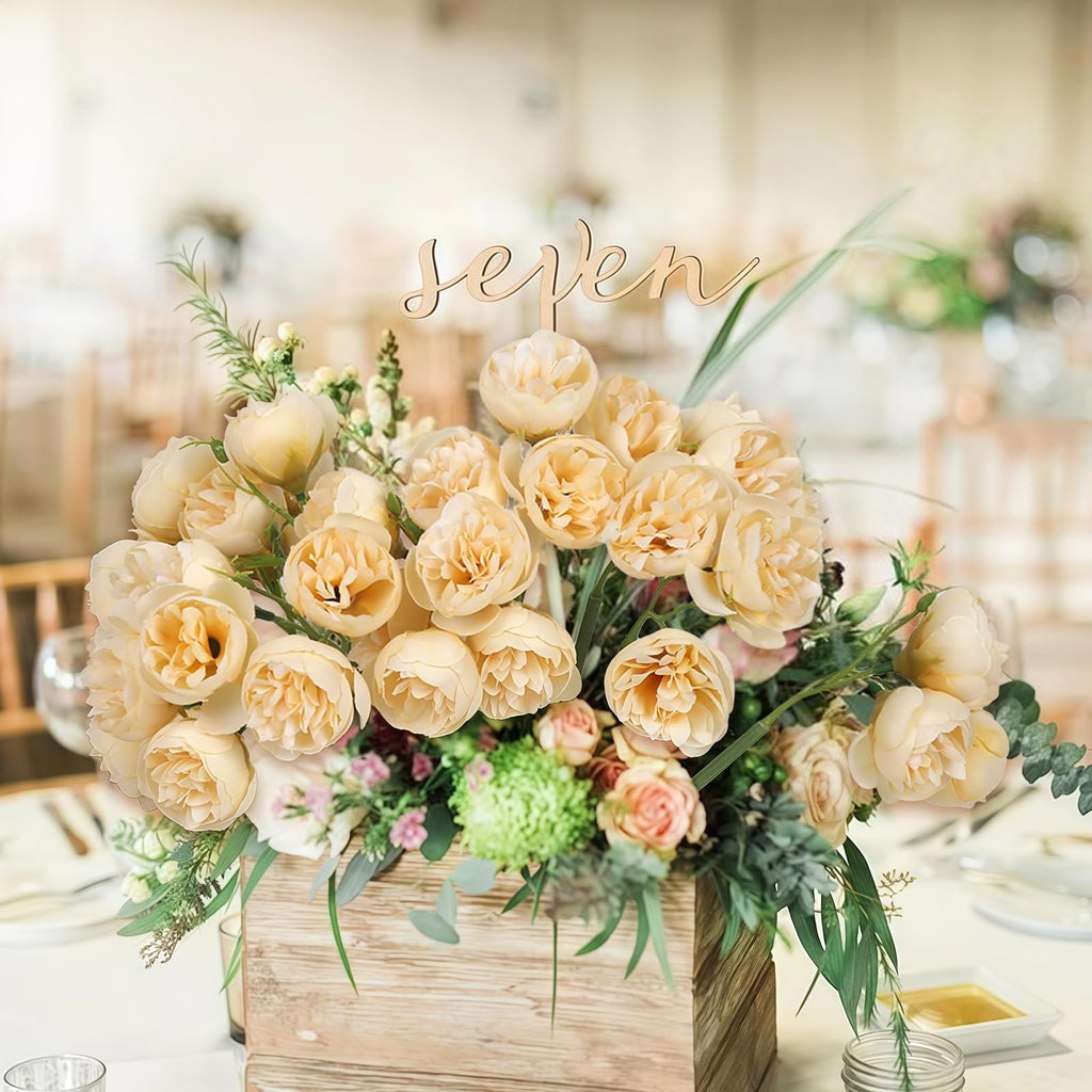 Champagne artificial peonies and ranunculus flowers in a wooden box with a 'seven' table number topper, set on a table.