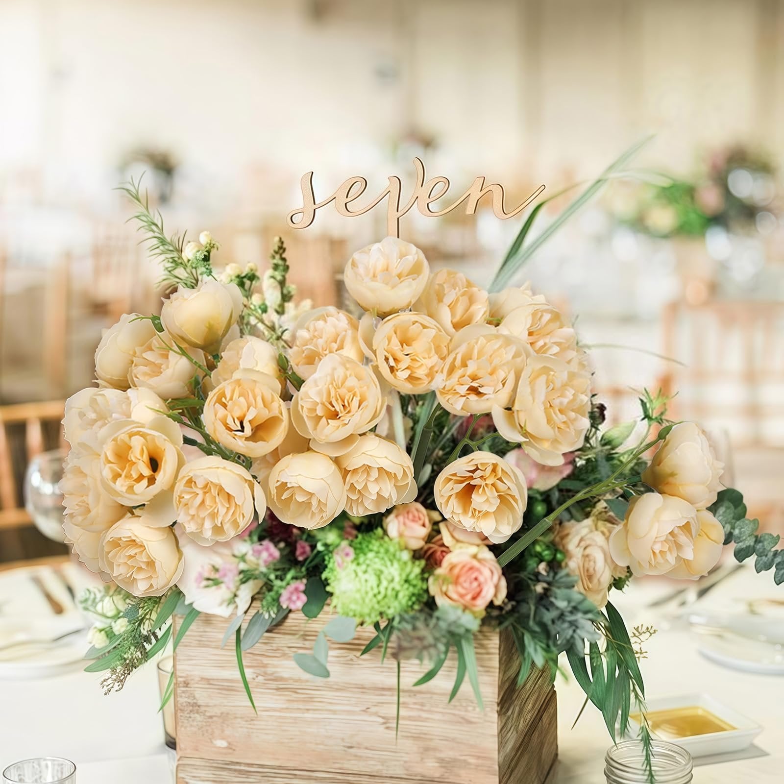 Champagne artificial peonies and ranunculus flowers in a wooden box with a 'seven' table number topper, set on a table.