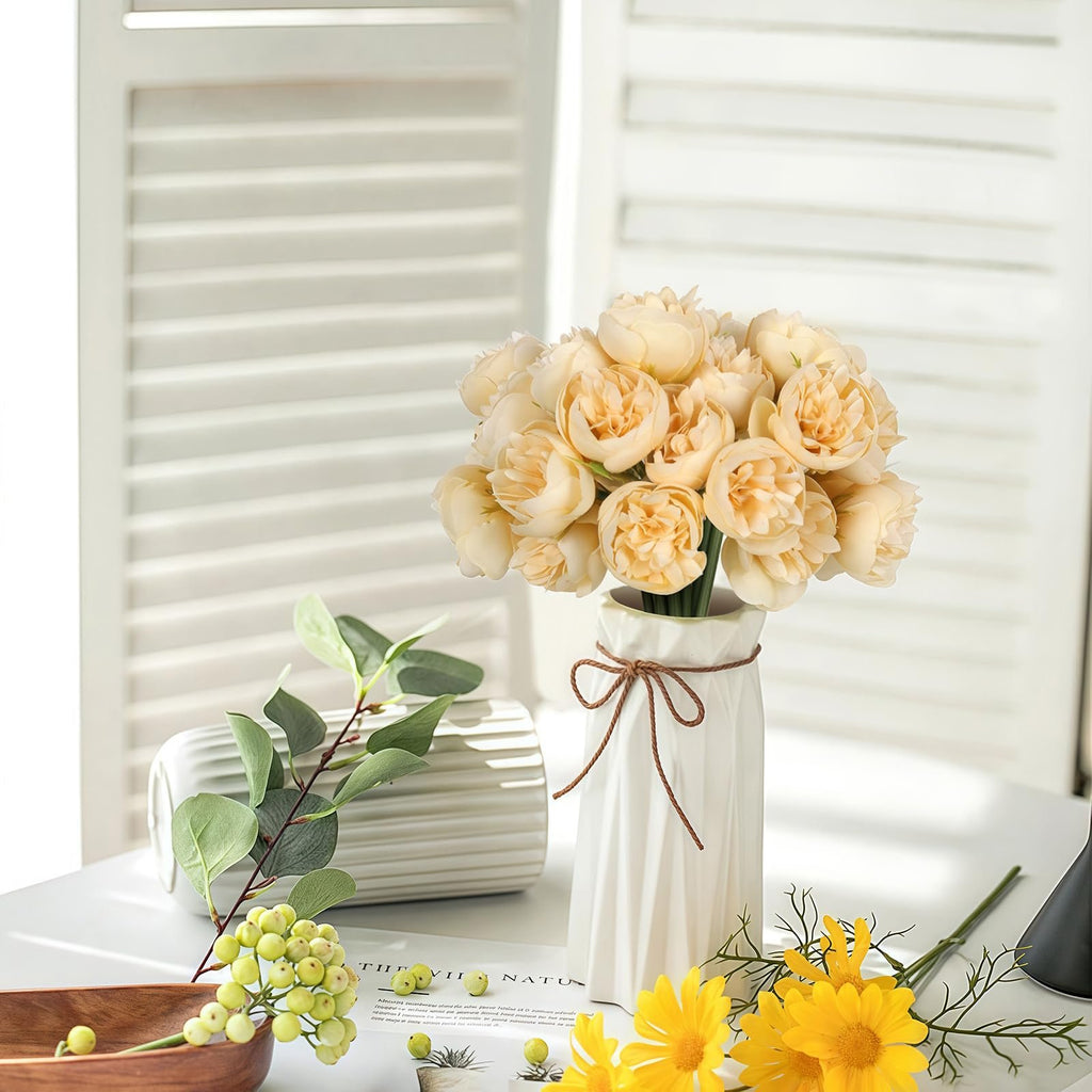 Champagne artificial peonies and ranunculus flowers in a white vase with decorative rope, next to eucalyptus branches and yellow daisies.