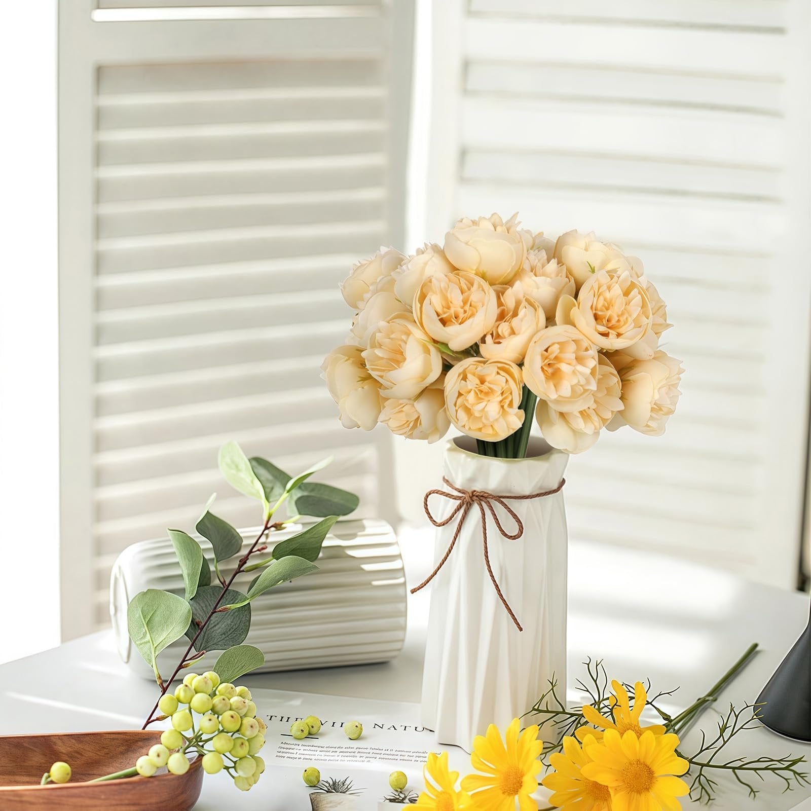 Champagne artificial peonies and ranunculus flowers in a white vase with decorative rope, next to eucalyptus branches and yellow daisies.