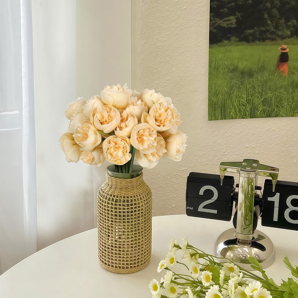 Champagne artificial peonies and ranunculus flowers in a woven vase on a table with a flip clock and daisies
