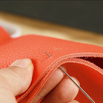 Close-up of a hand using a large-eye sewing needle to pierce orange leather material.