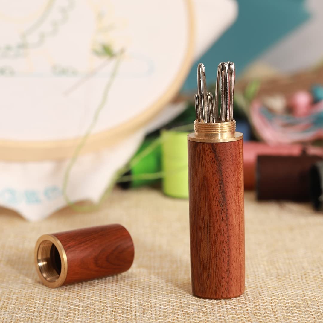 Wooden sewing needle case with brass accents, open with needles visible, and cap beside it. Embroidery hoop and threads in background.