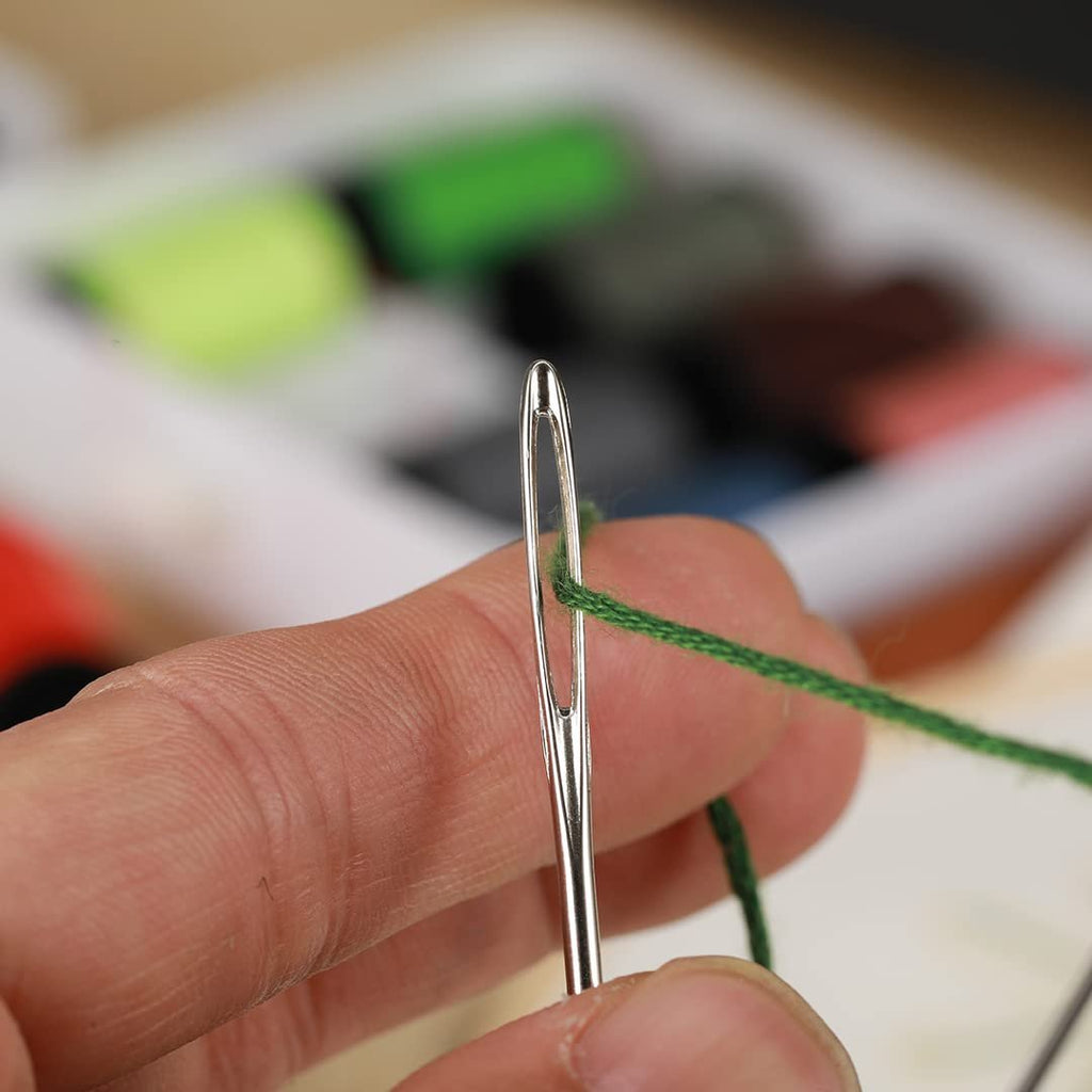 Close-up of a hand threading green yarn through the eye of a sewing needle. A sewing kit is blurred in the background.