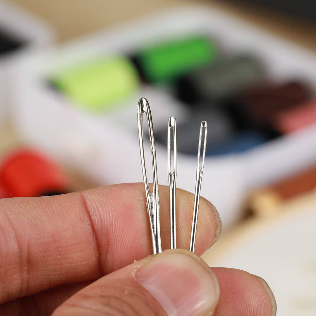 Close-up of three stainless steel sewing needles with large eyes held by a hand, with spools of thread blurred in the background.