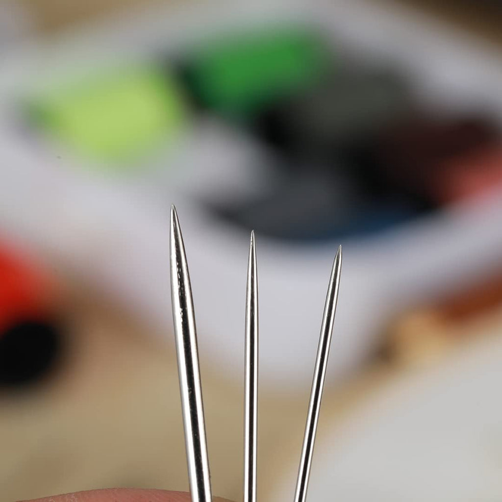 Close-up of three sewing needles with sharp points, showing varying thicknesses against a blurred background of colorful craft supplies.