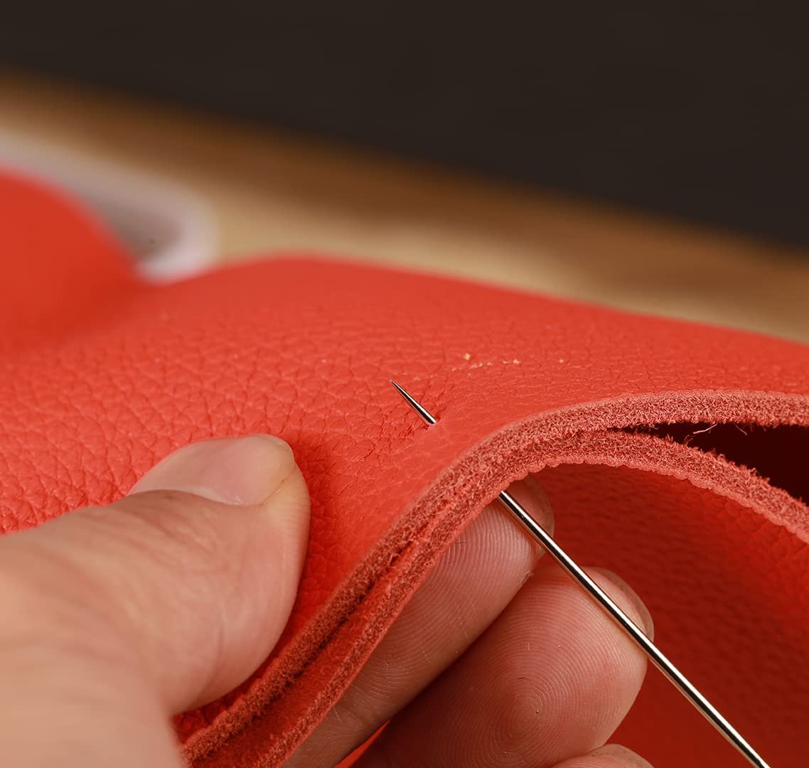 Close-up of a hand using a large-eye sewing needle to pierce red leather material.