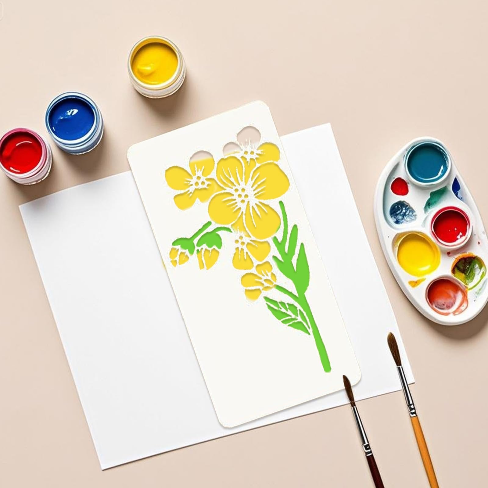 Flower stencil template with yellow flowers and green leaves, next to paints and brushes on paper.
