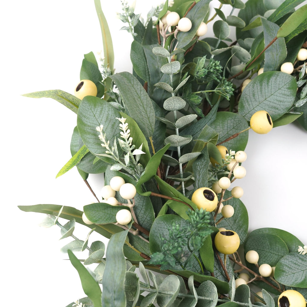 Close-up of an artificial olive and eucalyptus wreath with white berries and yellow accents on a white background.