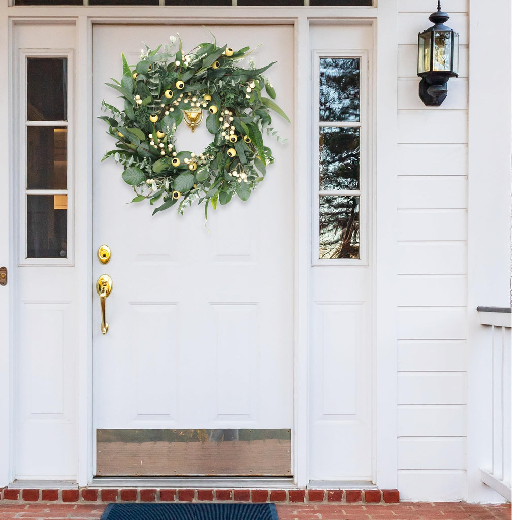 Artificial olive and eucalyptus wreath with white berries and gold accents hanging on a white front door.