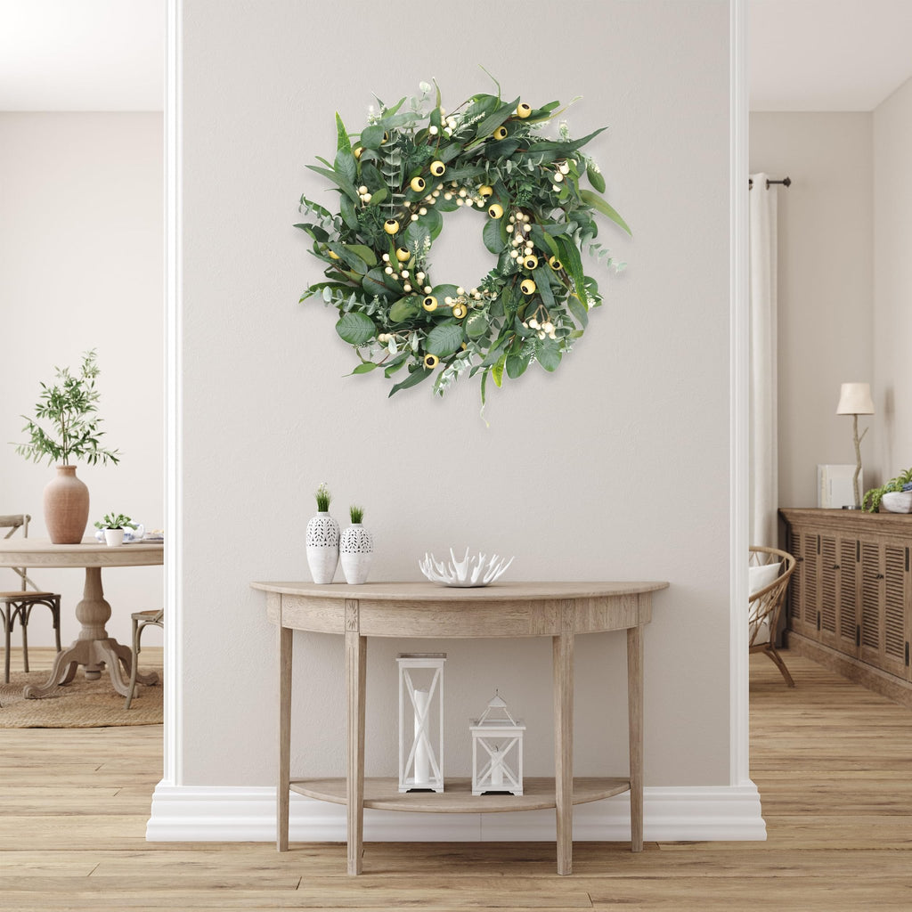 Artificial olive and eucalyptus wreath with white berries and gold lights hanging on a wall above a console table.