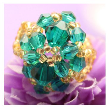 Close-up of a handmade ring crafted from faceted teal glass beads and small gold seed beads, resting on a purple flower petal.