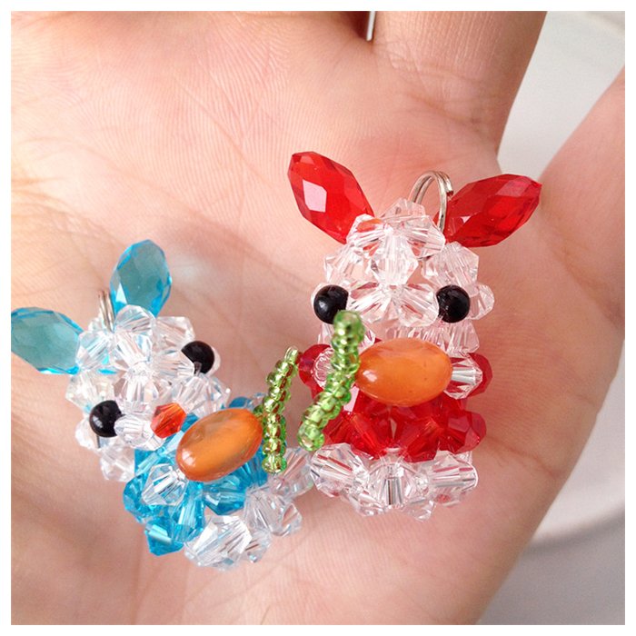Two beaded bunny charms, one blue and one red, held in a hand. The bunnies are made of clear, blue, and red faceted beads with orange carrot accents.