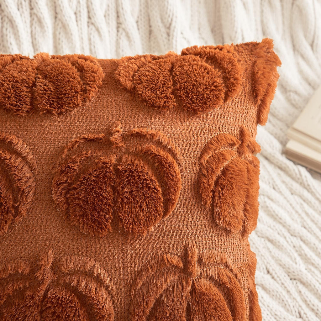 Close-up of a dark rust faux fur throw pillow cover with raised pumpkin designs, resting on a white cable-knit blanket.