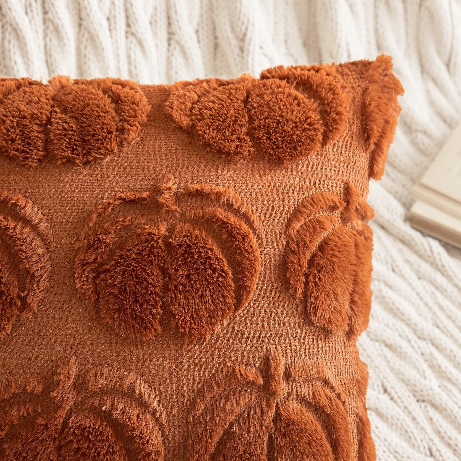 Close-up of a dark rust faux fur throw pillow cover with raised pumpkin designs, resting on a white cable-knit blanket.