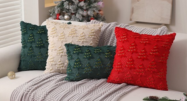 Four Christmas throw pillows with embroidered trees and lights on a couch with a Christmas tree in the background.