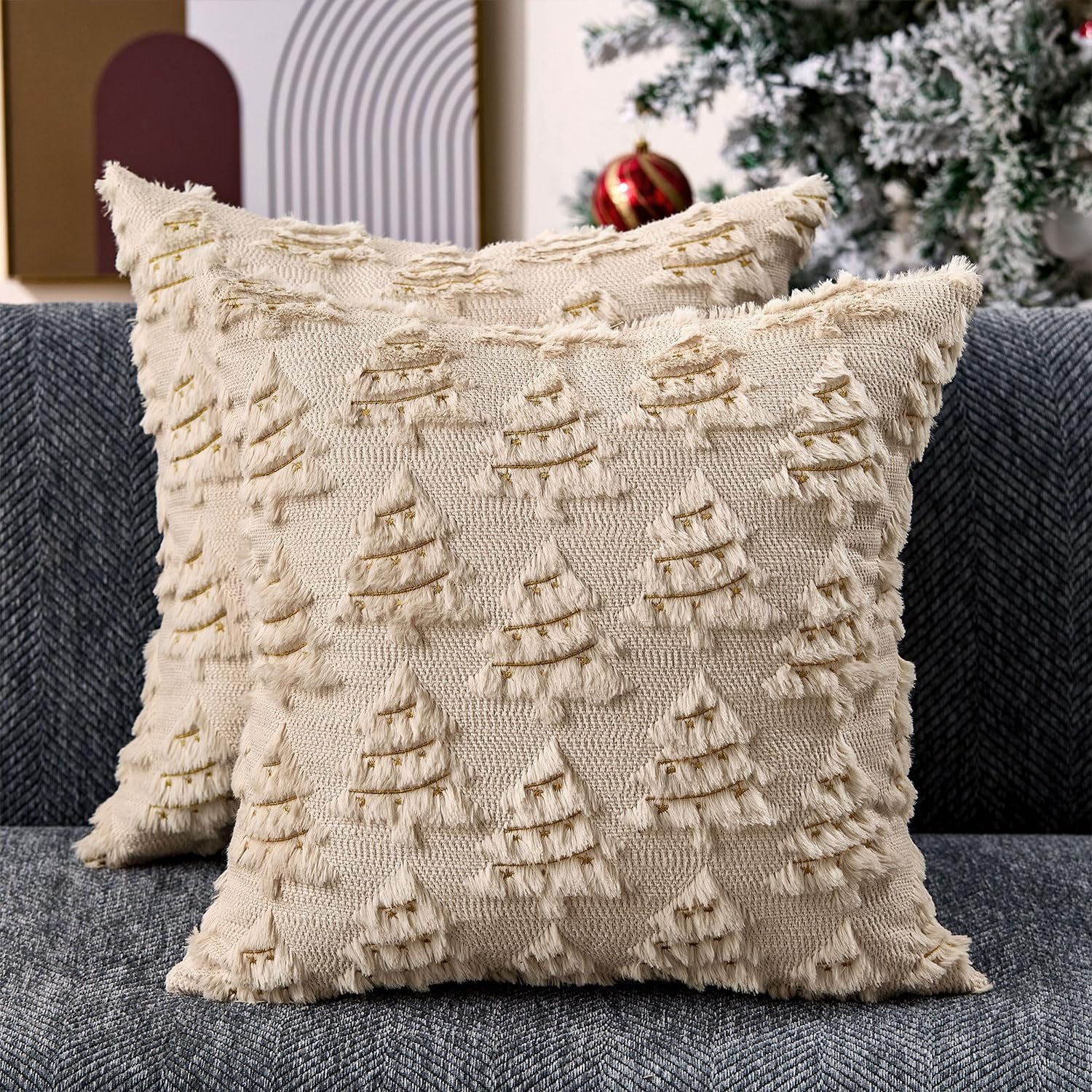 Two beige Christmas pillow covers with embroidered trees and gold accents on a sofa, with a Christmas tree in the background.