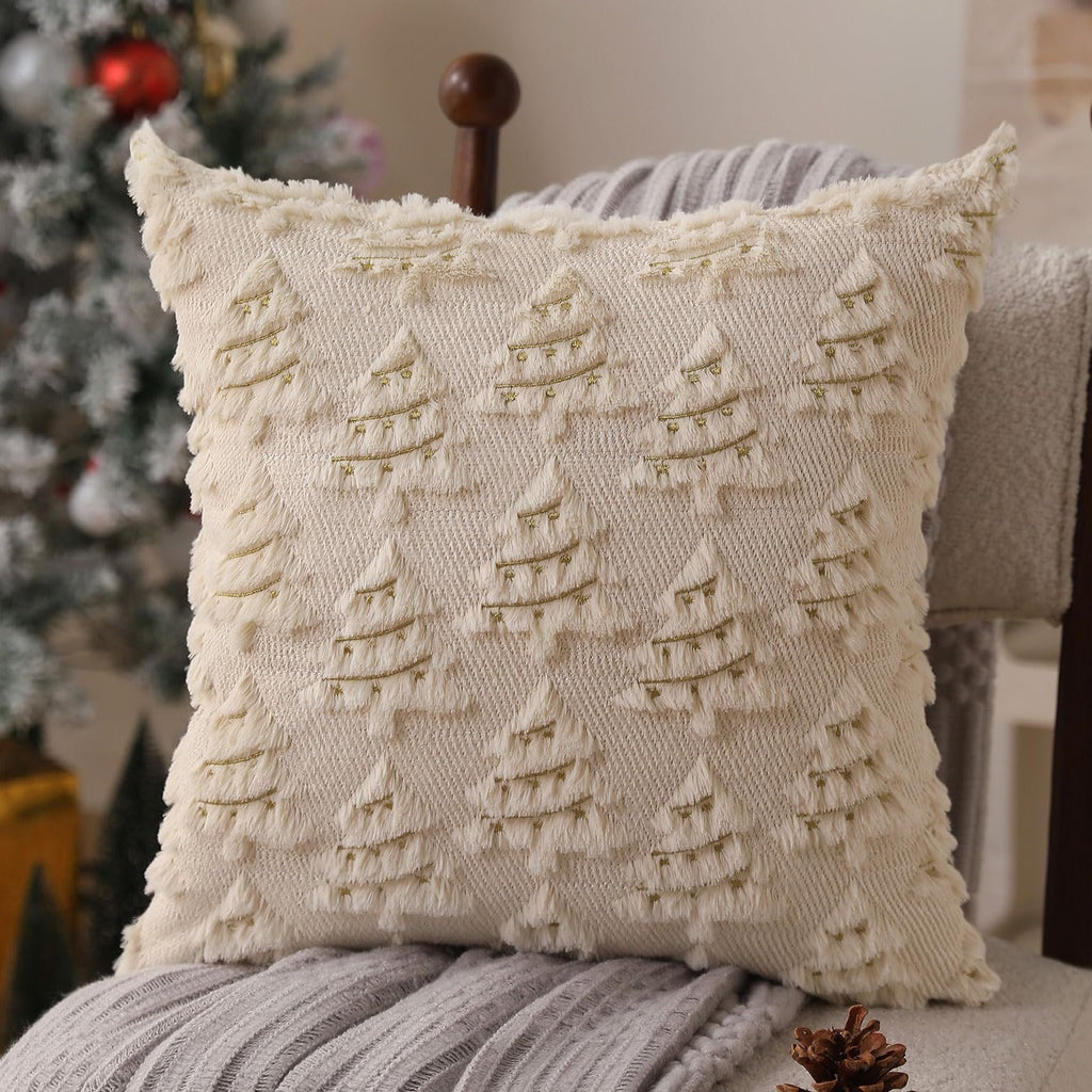 Beige Christmas pillow cover with embroidered trees and gold lights, on a chair with blankets and a Christmas tree in the background.