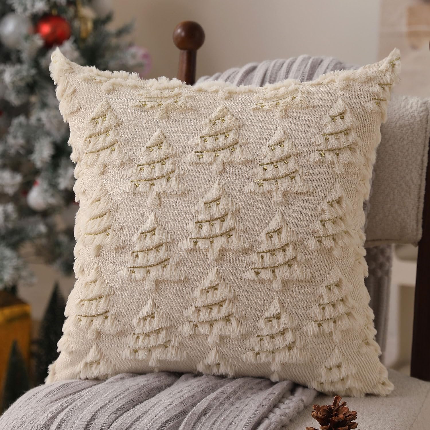 Beige Christmas pillow cover with embroidered trees and gold lights, on a chair with blankets and a Christmas tree in the background.