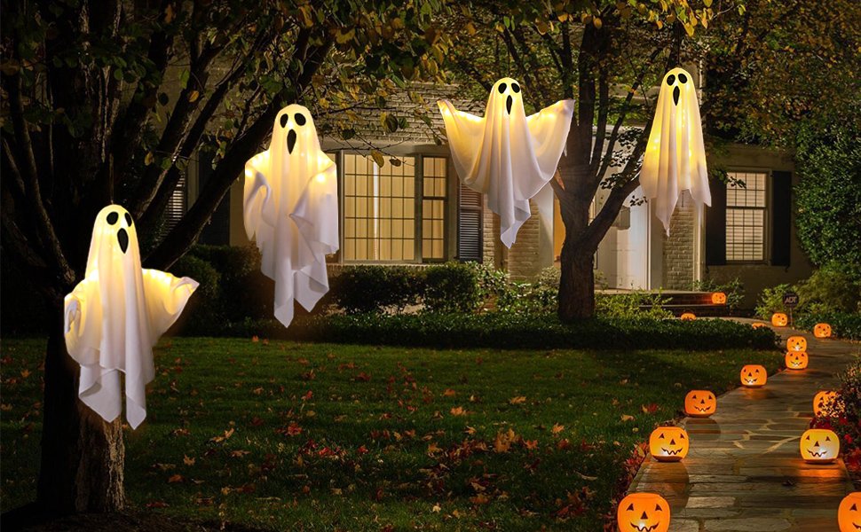 Four light-up hanging ghost decorations suspended from trees in a yard with illuminated jack-o'-lanterns lining a pathway.