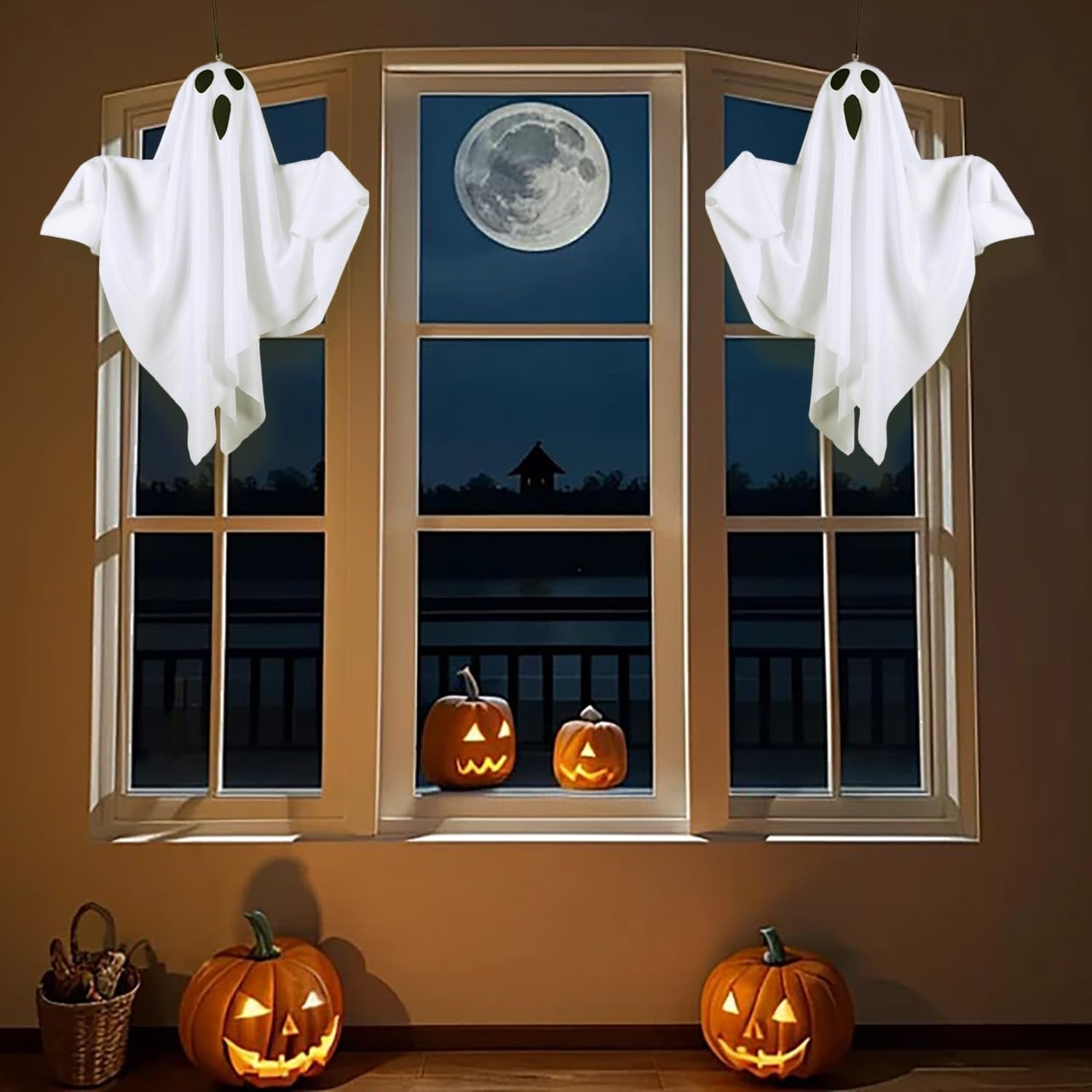 Two hanging white ghost decorations with black eyes and mouths, illuminated by warm white LEDs, displayed in a window with carved pumpkins below.