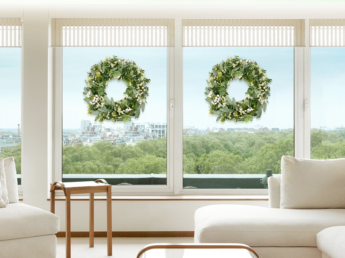 Two artificial green eucalyptus wreaths with boxwood and white berries displayed on a large window overlooking a city and forest.