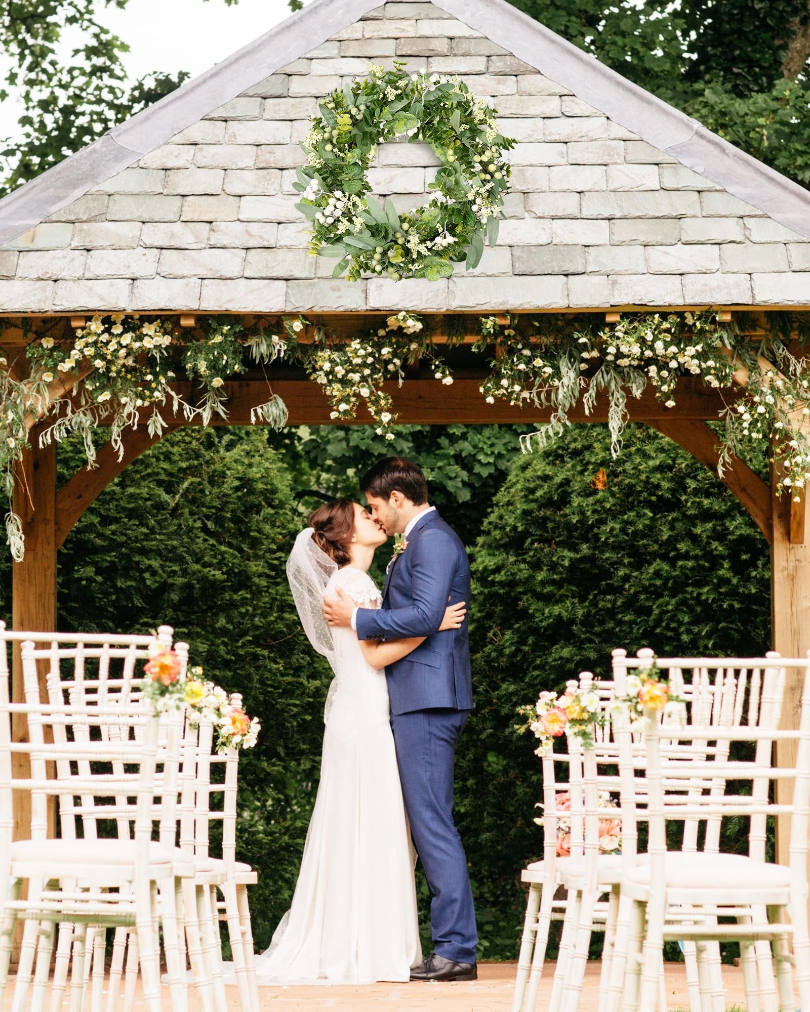 Wedding couple kissing under a gazebo decorated with a green eucalyptus wreath and floral arrangements.