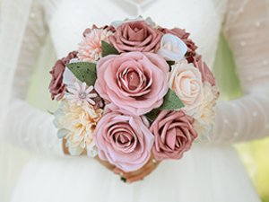 Dusty blush pink artificial rose bouquet with various flowers and green leaves held by a person in a white wedding dress.
