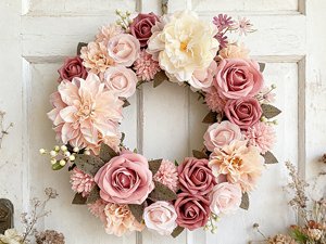 Dusty blush pink artificial rose and dahlia floral wreath with green leaves, displayed on a white wooden door.