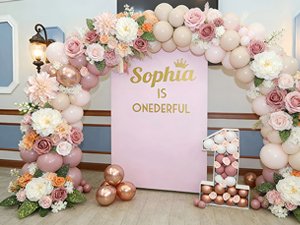 Birthday party decor with a pink backdrop, balloons, and floral arrangements, featuring a large number '1' and the text 'Sophia is onederful'.