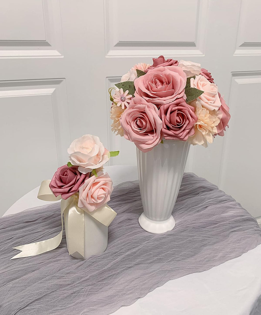 Two arrangements of dusty blush pink artificial roses in white vases, one larger bouquet in a fluted vase, and a smaller one tied with a cream ribbon.
