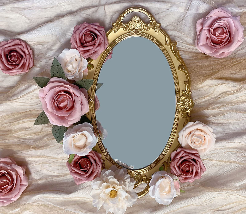 Oval gold ornate mirror surrounded by dusty blush pink and cream artificial roses on a textured cream fabric.