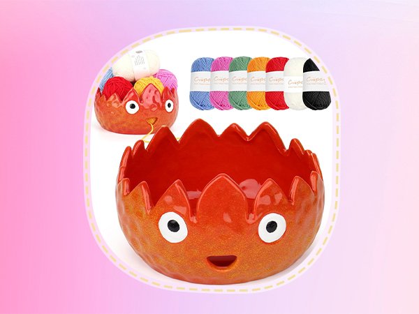 Two orange ceramic yarn bowls with faces, one filled with yarn balls and the other empty, with a row of colorful yarn skeins behind.