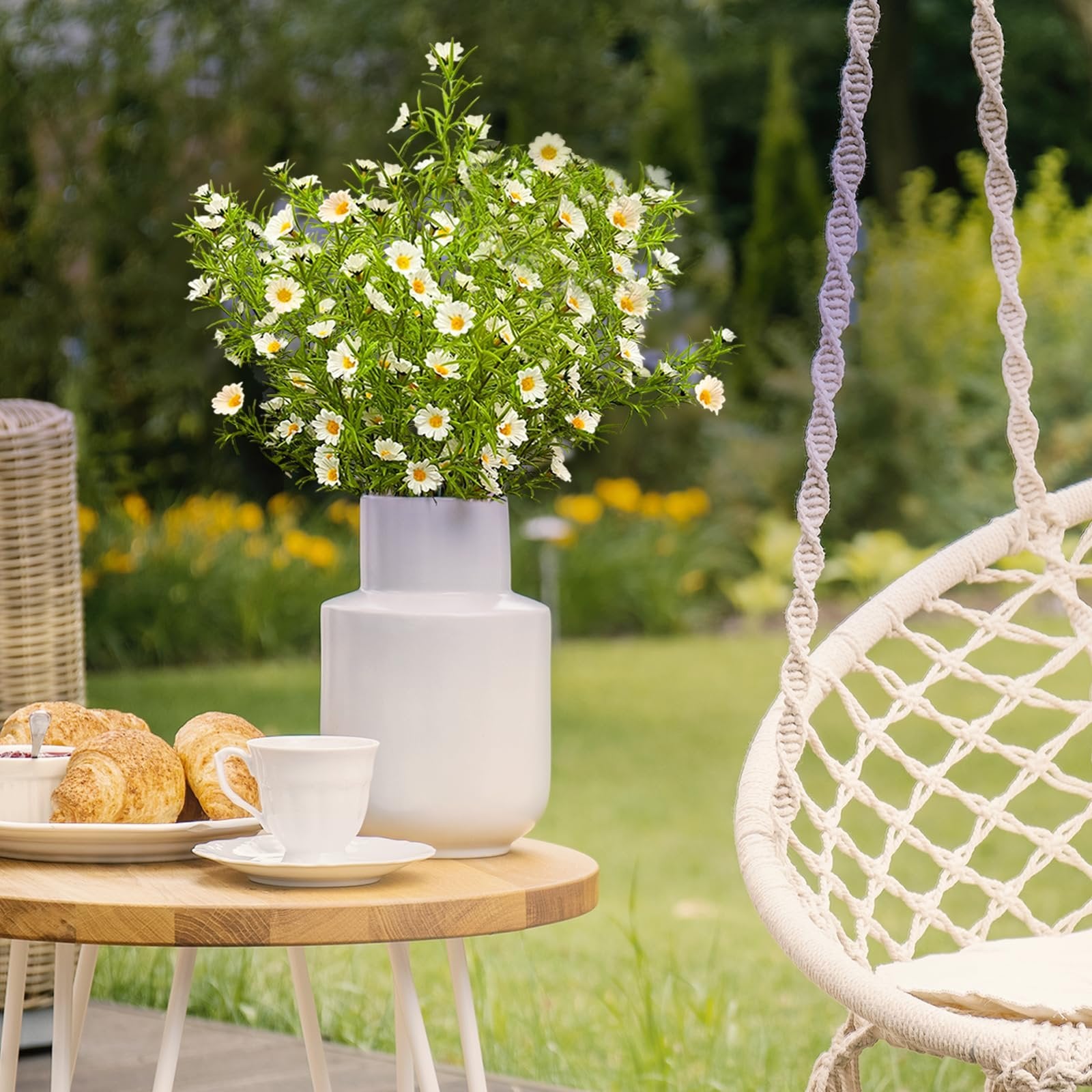 Artificial white daisy flowers in a vase on a table with croissants and coffee cup, next to a hanging chair outdoors.