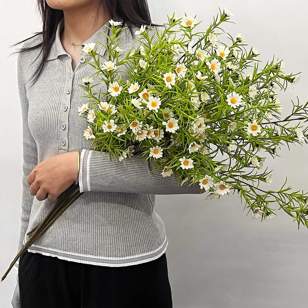 Person holding a bouquet of artificial white daisy flowers with green stems and leaves against a white background.