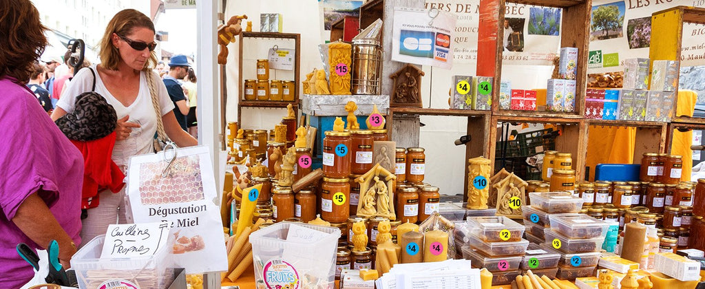 Outdoor market stall with jars of honey and beeswax candles, a woman browsing.