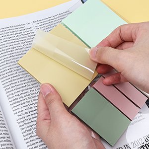 Hands peeling a yellow transparent sticky note from a stack of multicolor sticky notes on a newspaper.