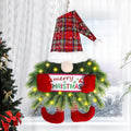 Christmas gnome wreath with plaid hat, pine branches, lights, and a 'Merry Christmas' banner, hanging indoors.