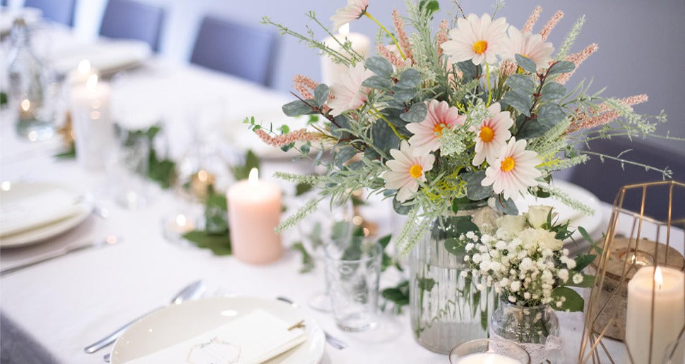 Artificial wildflower bouquet with pink daisies and eucalyptus in a glass vase on a decorated table with candles.