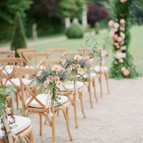 Artificial wildflower bouquet with pink daisies and eucalyptus tied to a wooden chair at an outdoor event.
