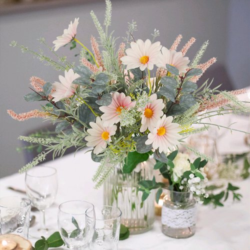 Two artificial wildflower bouquets with pink daisies and eucalyptus in a glass vase on a table setting.