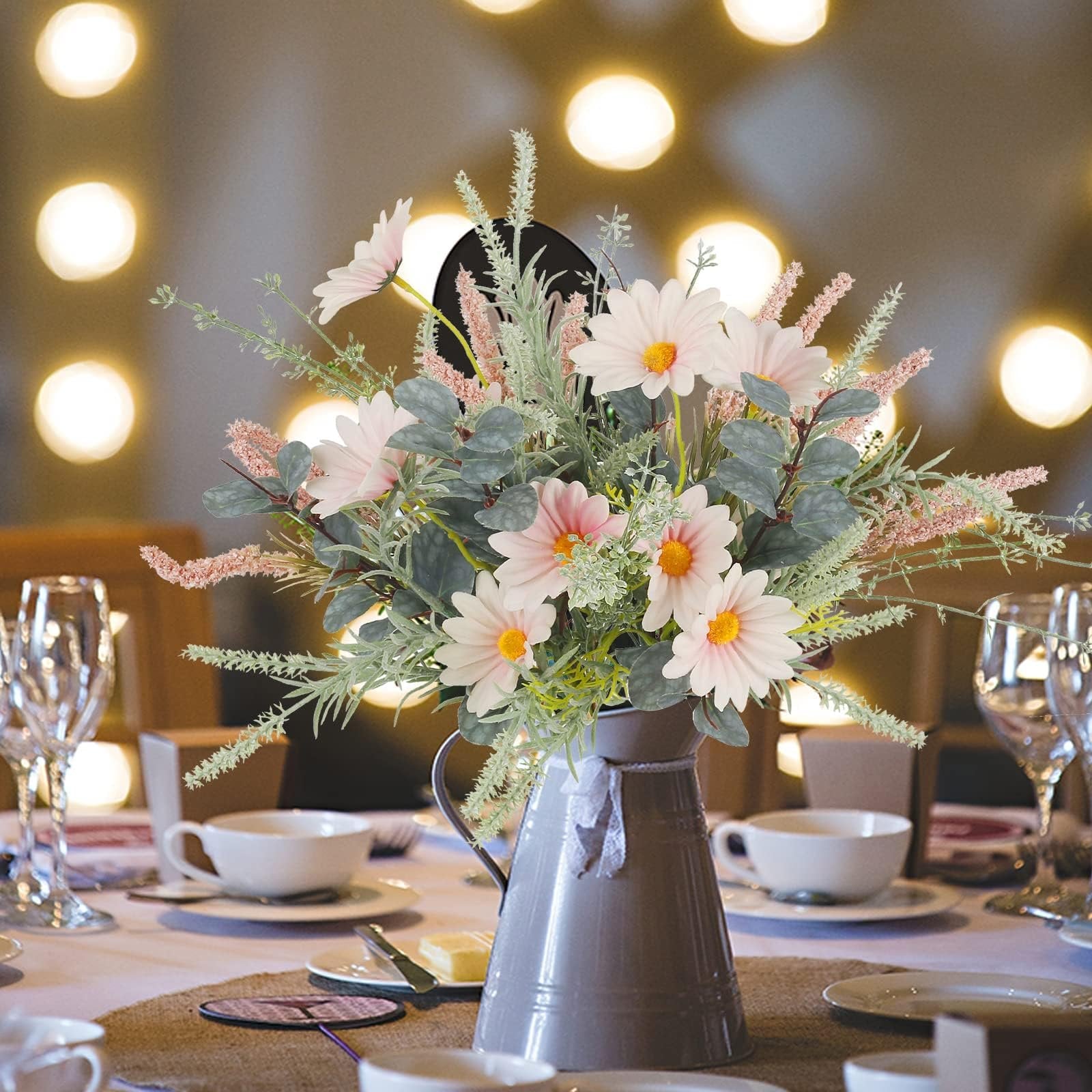 Artificial wildflower bouquet with pink daisies and eucalyptus in a gray pitcher on a table setting with bokeh lights