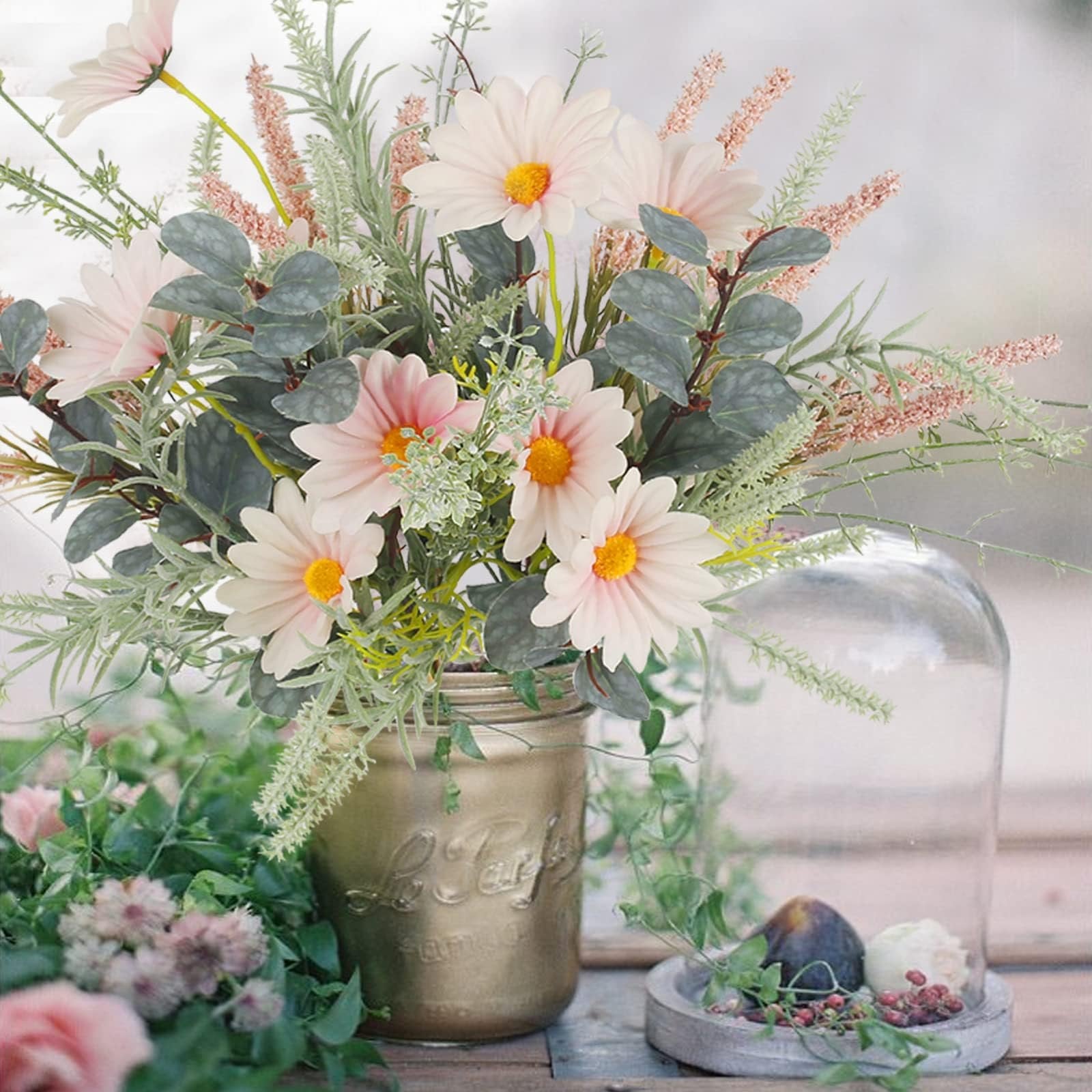 Artificial wildflower bouquet with pink daisies and eucalyptus in a gold mason jar, next to a glass cloche with figs.